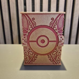 Peut inclure: Une bo&icirc;te rose et dor&eacute;e orn&eacute;e d'un motif de Pok&eacute; Ball au centre. La bo&icirc;te pr&eacute;sente des motifs et des dessins complexes sur ses c&ocirc;t&eacute;s et ses bords. Elle repose sur une surface gris clair.