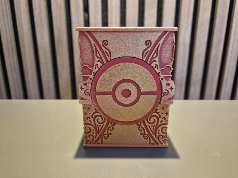 Peut inclure: Une bo&icirc;te rose et dor&eacute;e orn&eacute;e d'un motif de Pok&eacute; Ball au centre. La bo&icirc;te pr&eacute;sente des motifs et des dessins complexes sur ses c&ocirc;t&eacute;s et ses bords. Elle repose sur une surface gris clair.
