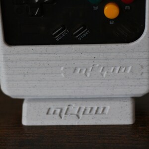 May include: A white handheld gaming console with black buttons and a speckled finish. The console has a black and white screen and the words "retro-bit" printed on the front. The console is sitting on a white stand with the word "retro-bit" printed on the front.