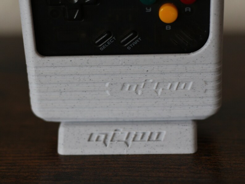 May include: A white handheld gaming console with black buttons and a speckled finish. The console has a black and white screen and the words "retro-bit" printed on the front. The console is sitting on a white stand with the word "retro-bit" printed on the front.