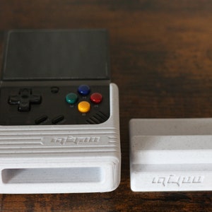 May include: A white handheld gaming console with a black screen and buttons. The console has a stand that can be used to prop it up. The console has the word "Anbernic" printed on it.