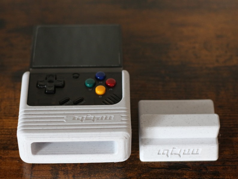 May include: A white handheld gaming console with a black screen and buttons. The console has a stand that can be used to prop it up. The console has the word "Anbernic" printed on it.
