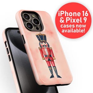 Puede incluir: Funda de teléfono rosa claro con una ilustración de acuarela de un soldado cascanueces con uniforme rojo y sombrero negro. La funda se muestra con un teléfono negro. Un círculo rojo indica "iPhone 16 & Pixel 9 cases now available!"