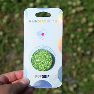 May include: A green glitter PopSocket PopGrip phone grip. The packaging features the PopSockets logo and the text "Try me! Pull twice to grip".