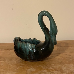 May include: A ceramic swan-shaped planter with a glossy, dark teal and black glaze. The swan has a curved neck and a scalloped edge on the planter's opening. The planter is sitting on a wooden surface.