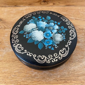 May include: A black round trinket box with a gold floral design. The flowers are white and blue with a black background.