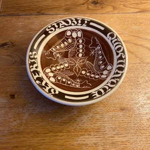 May include: A white ceramic bowl with a brown glaze and a white design of three stylised butterflies. The bowl has the words "Stabit Quotidie" written around the rim.