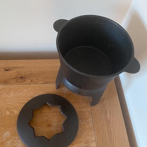 May include: A black cast iron pot with three legs and a matching lid with a star-shaped cutout. The pot is sitting on a wooden surface.