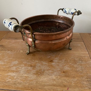 May include: A copper planter with two white ceramic handles with blue floral designs. The planter has three brass legs and is sitting on a wooden surface.