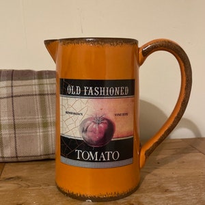 May include: Orange ceramic jug with a brown handle. The jug has a label that says "Old Fashioned Homegrown Vine Ripe Tomato".