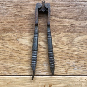 May include: A vintage, black metal nutcracker with a simple design. The nutcracker has two long handles with a textured grip and a hinged jaw in the centre.