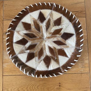 May include: A round, brown and white leather patchwork rug with a starburst design. The rug is stitched together with white thread and has a white leather border.