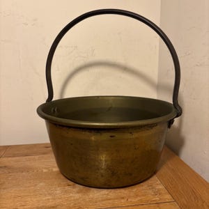 May include: An antique brass cauldron with a dark metal handle. The cauldron has a rounded body and a wide opening. The handle arches over the top, attached on either side. The brass has a warm, aged patina, suggesting its use over time.