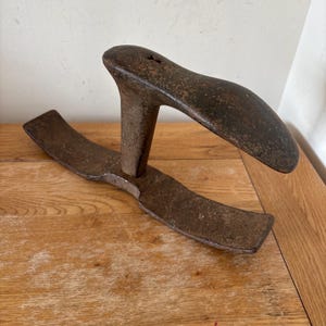 May include: A vintage, weathered metal shoe last, featuring a curved shoe form atop a T-shaped base. The metal has a dark, aged patina, suggesting years of use. The shoe form is approximately 25 cm long.