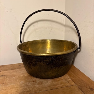May include: An antique brass cauldron, featuring a dark, aged exterior and a polished, golden interior. The cauldron has a rounded form and a strong, arched handle made of dark metal. It rests on a wooden surface, evoking a rustic or vintage feel.