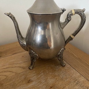 May include: A silver teapot with a decorative handle and three feet. The teapot has a round body and a domed lid.