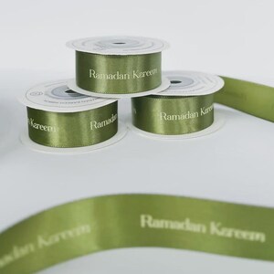 May include: Olive green ribbon rolls and loose ribbon with the text "Ramadan Kareem" printed in white. Three rolls are stacked, with additional ribbon pieces scattered around.