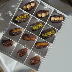 May include: A white box divided into sections, each containing a date. Some dates are filled with nuts, others with a green filling, and some with a yellow filling. The dates are dark brown, and the box is open.
