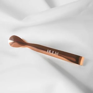 May include: A copper-colored ice tong with a rounded scoop end and a flat, rectangular handle. The word "HULM" is printed in white on the handle. The tong is set against a white, textured background.
