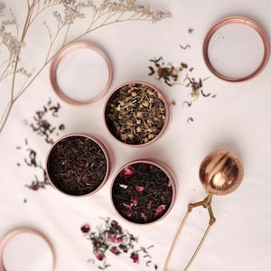 May include: Four rose gold metal tea tins with different types of loose leaf tea. The tins are on a white surface with a rose gold tea infuser.
