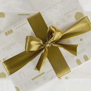 May include: A wrapped gift, presented in white paper with a repeating gold architectural design. It is adorned with a wide, gold satin ribbon and a large bow. The overall appearance is elegant and celebratory.