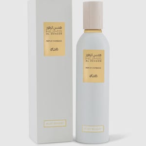 May include: A white perfume bottle with a wooden cap and a matching box. The bottle and box have gold labels with Arabic and French text. The text on the bottle reads "HUMS AL ZOHOOR PARFUM D'AMBIANCE" and "VELVET BOUQUET".