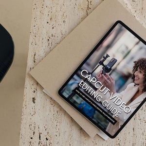 May include: A tablet displaying a guide on how to use CapCut for video editing. The guide is titled "CapCut Video Editing Guide: How to Use CapCut & Video Editing Walkthroughs". The image shows a person holding a smartphone and a tripod.