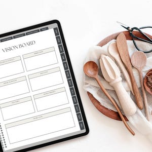 May include: A digital tablet displaying a vision board planner page with sections for goals, health, and self-love, alongside a wooden bowl containing a light-colored fabric, wooden kitchen utensils, a citrus reamer, and black metal scissors.