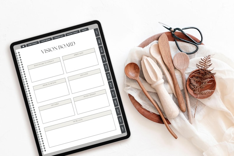 May include: A digital tablet displaying a vision board planner page with sections for goals, health, and self-love, alongside a wooden bowl containing a light-colored fabric, wooden kitchen utensils, a citrus reamer, and black metal scissors.