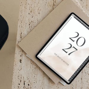 May include: A digital tablet displaying a 2027 planner with a beige watercolor background. The screen shows the text 2027 and THE BEST YEAR EVER in a serif font. The tablet features a black frame and a side navigation bar with monthly tabs.