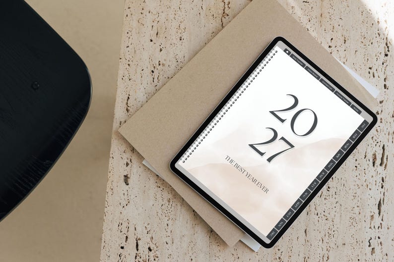 May include: A digital tablet displaying a 2027 planner with a beige watercolor background. The screen shows the text 2027 and THE BEST YEAR EVER in a serif font. The tablet features a black frame and a side navigation bar with monthly tabs.
