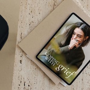 May include: A tablet computer displaying a digital magazine cover with the title "Coping with grief". The cover features a photo of a person with their hand on their face, looking down.
