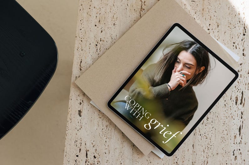 May include: A tablet computer displaying a digital magazine cover with the title "Coping with grief". The cover features a photo of a person with their hand on their face, looking down.