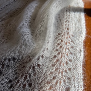 Elbert is a delicate, hand-knitted lace shawl made from soft, white mohair/silk yarn. The intricate lace pattern features openwork stitches, creating an airy and lightweight texture. The scalloped edges add elegance, making this piece perfect for formal occasions or as a cozy, stylish accessory. The natural light highlights the fine, haloed fibers, enhancing the ethereal quality of the knitwear. Ideal for those searching for luxury handmade shawls, bridal wraps, or heirloom-quality knitwear
