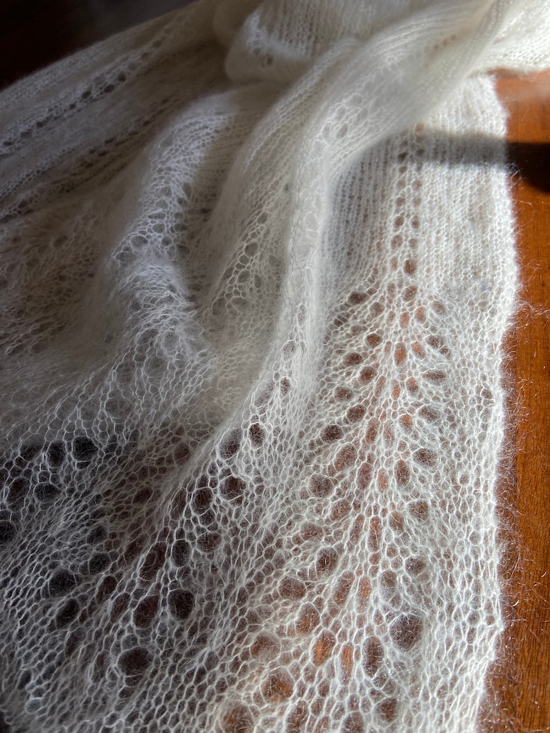 Elbert is a delicate, hand-knitted lace shawl made from soft, white mohair/silk yarn. The intricate lace pattern features openwork stitches, creating an airy and lightweight texture. The scalloped edges add elegance, making this piece perfect for formal occasions or as a cozy, stylish accessory. The natural light highlights the fine, haloed fibers, enhancing the ethereal quality of the knitwear. Ideal for those searching for luxury handmade shawls, bridal wraps, or heirloom-quality knitwear