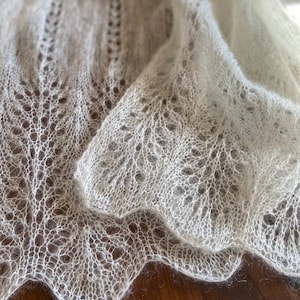 Elbert is a delicate, hand-knitted lace shawl made from soft, white mohair/silk yarn. The intricate lace pattern features openwork stitches, creating an airy and lightweight texture. The scalloped edges add elegance, making this piece perfect for formal occasions or as a cozy, stylish accessory. The natural light highlights the fine, haloed fibers, enhancing the ethereal quality of the knitwear. Ideal for those searching for luxury handmade shawls, bridal wraps, or heirloom-quality knitwear