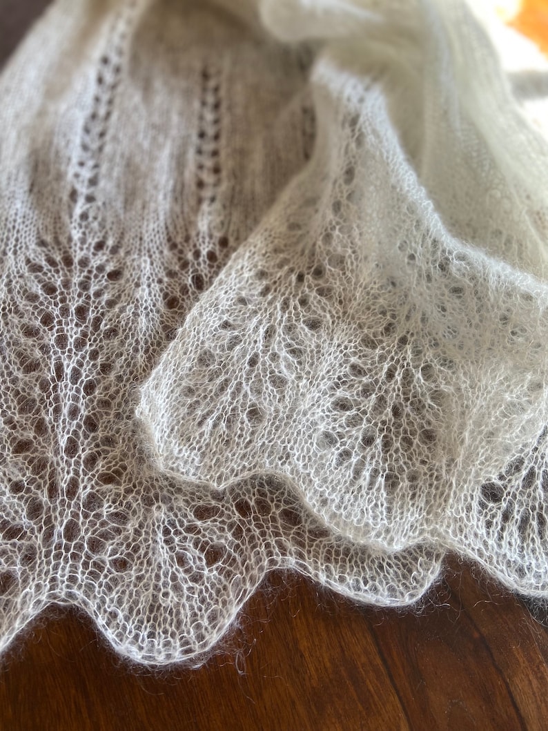 Elbert is a delicate, hand-knitted lace shawl made from soft, white mohair/silk yarn. The intricate lace pattern features openwork stitches, creating an airy and lightweight texture. The scalloped edges add elegance, making this piece perfect for formal occasions or as a cozy, stylish accessory. The natural light highlights the fine, haloed fibers, enhancing the ethereal quality of the knitwear. Ideal for those searching for luxury handmade shawls, bridal wraps, or heirloom-quality knitwear