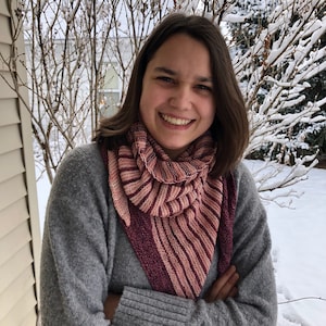 May include: A woman wearing a gray sweater with a pink and burgundy striped shawl. She is smiling and has her arms crossed. The background is a snowy landscape.