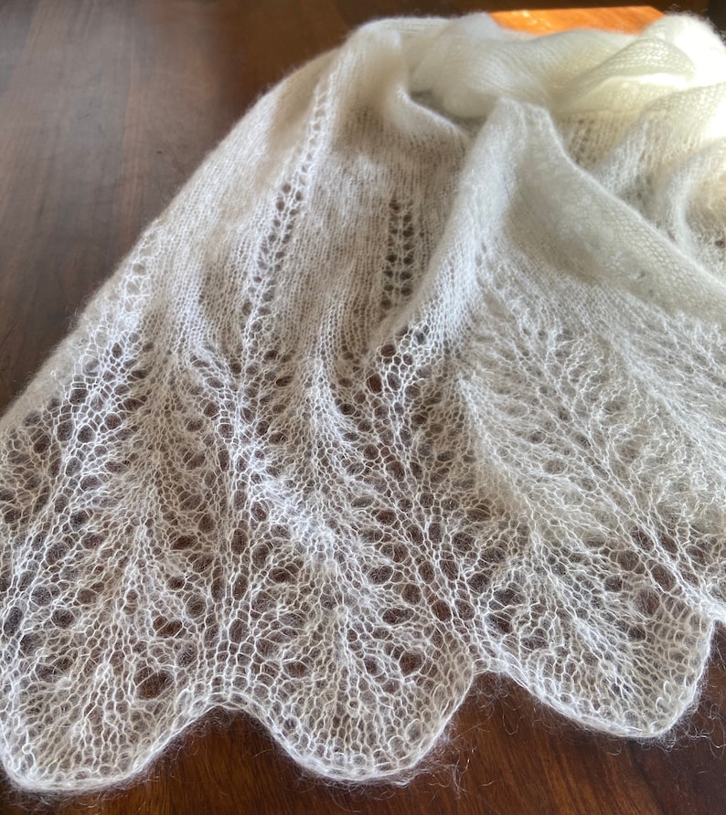 Elbert is a delicate, hand-knitted lace shawl made from soft, white mohair/silk yarn. The intricate lace pattern features openwork stitches, creating an airy and lightweight texture. The scalloped edges add elegance, making this piece perfect for formal occasions or as a cozy, stylish accessory. The natural light highlights the fine, haloed fibers, enhancing the ethereal quality of the knitwear. Ideal for those searching for luxury handmade shawls, bridal wraps, or heirloom-quality knitwear