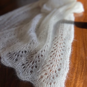 Elbert is a delicate, hand-knitted lace shawl made from soft, white mohair/silk yarn. The intricate lace pattern features openwork stitches, creating an airy and lightweight texture. The scalloped edges add elegance, making this piece perfect for formal occasions or as a cozy, stylish accessory. The natural light highlights the fine, haloed fibers, enhancing the ethereal quality of the knitwear. Ideal for those searching for luxury handmade shawls, bridal wraps, or heirloom-quality knitwear