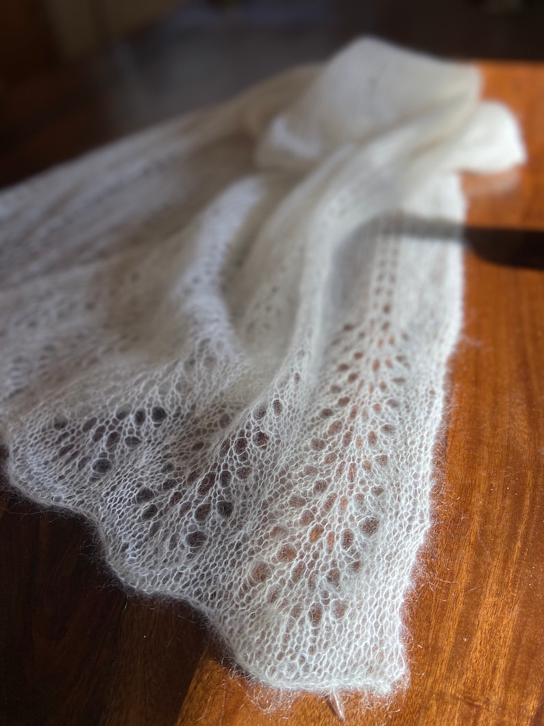 Elbert is a delicate, hand-knitted lace shawl made from soft, white mohair/silk yarn. The intricate lace pattern features openwork stitches, creating an airy and lightweight texture. The scalloped edges add elegance, making this piece perfect for formal occasions or as a cozy, stylish accessory. The natural light highlights the fine, haloed fibers, enhancing the ethereal quality of the knitwear. Ideal for those searching for luxury handmade shawls, bridal wraps, or heirloom-quality knitwear