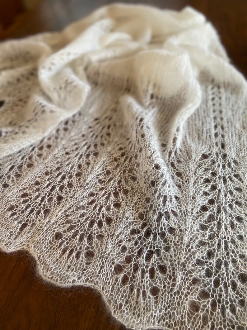 Elbert is a delicate, hand-knitted lace shawl made from soft, white mohair/silk yarn. The intricate lace pattern features openwork stitches, creating an airy and lightweight texture. The scalloped edges add elegance, making this piece perfect for formal occasions or as a cozy, stylish accessory. The natural light highlights the fine, haloed fibers, enhancing the ethereal quality of the knitwear. Ideal for those searching for luxury handmade shawls, bridal wraps, or heirloom-quality knitwear