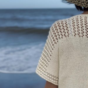 Puede incluir: Primer plano de una mujer con un top blanco de ganchillo con un delicado patrón de encaje. El top tiene mangas cortas y un corte holgado. La mujer está de pie frente a un fondo borroso de una playa y el océano.