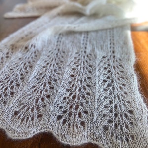 Elbert is a delicate, hand-knitted lace shawl made from soft, white mohair/silk yarn. The intricate lace pattern features openwork stitches, creating an airy and lightweight texture. The scalloped edges add elegance, making this piece perfect for formal occasions or as a cozy, stylish accessory. The natural light highlights the fine, haloed fibers, enhancing the ethereal quality of the knitwear. Ideal for those searching for luxury handmade shawls, bridal wraps, or heirloom-quality knitwear