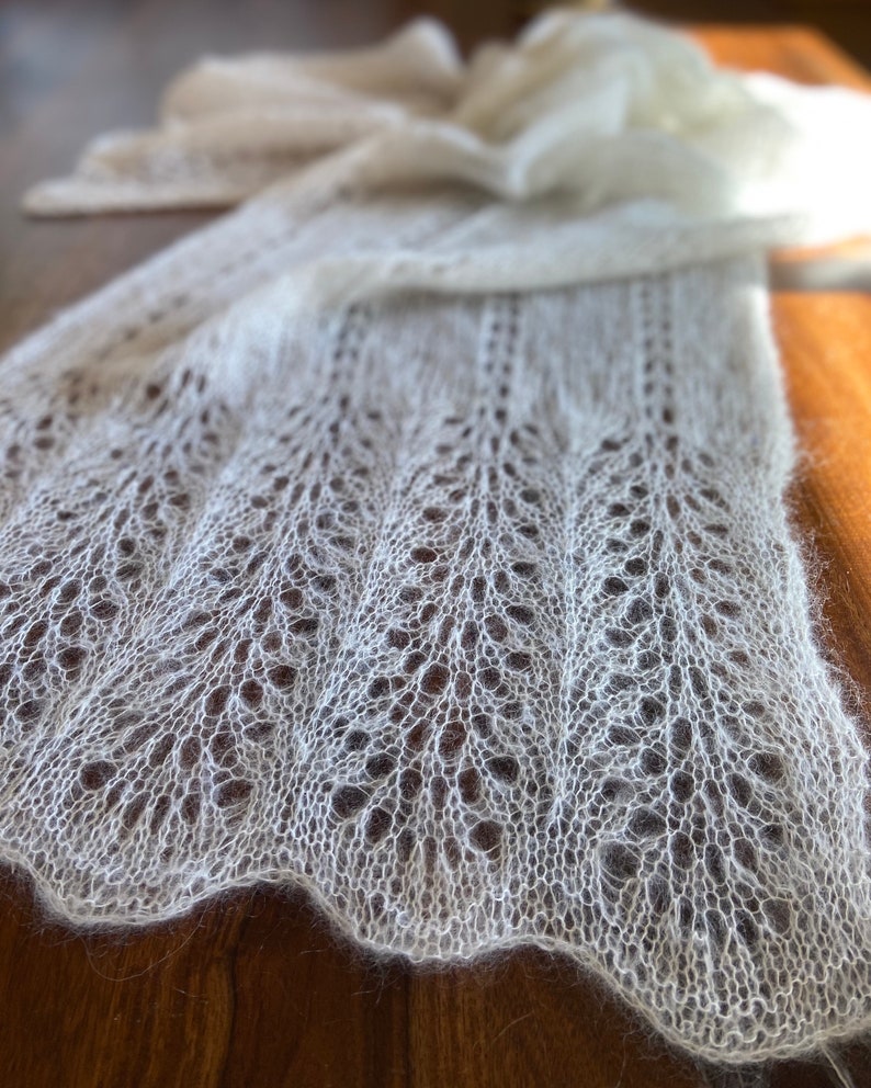 Elbert is a delicate, hand-knitted lace shawl made from soft, white mohair/silk yarn. The intricate lace pattern features openwork stitches, creating an airy and lightweight texture. The scalloped edges add elegance, making this piece perfect for formal occasions or as a cozy, stylish accessory. The natural light highlights the fine, haloed fibers, enhancing the ethereal quality of the knitwear. Ideal for those searching for luxury handmade shawls, bridal wraps, or heirloom-quality knitwear