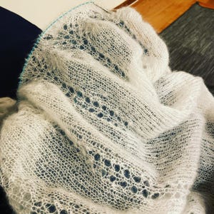 Elbert is a delicate, hand-knitted lace shawl made from soft, white mohair/silk yarn. The intricate lace pattern features openwork stitches, creating an airy and lightweight texture. The scalloped edges add elegance, making this piece perfect for formal occasions or as a cozy, stylish accessory. The natural light highlights the fine, haloed fibers, enhancing the ethereal quality of the knitwear. Ideal for those searching for luxury handmade shawls, bridal wraps, or heirloom-quality knitwear