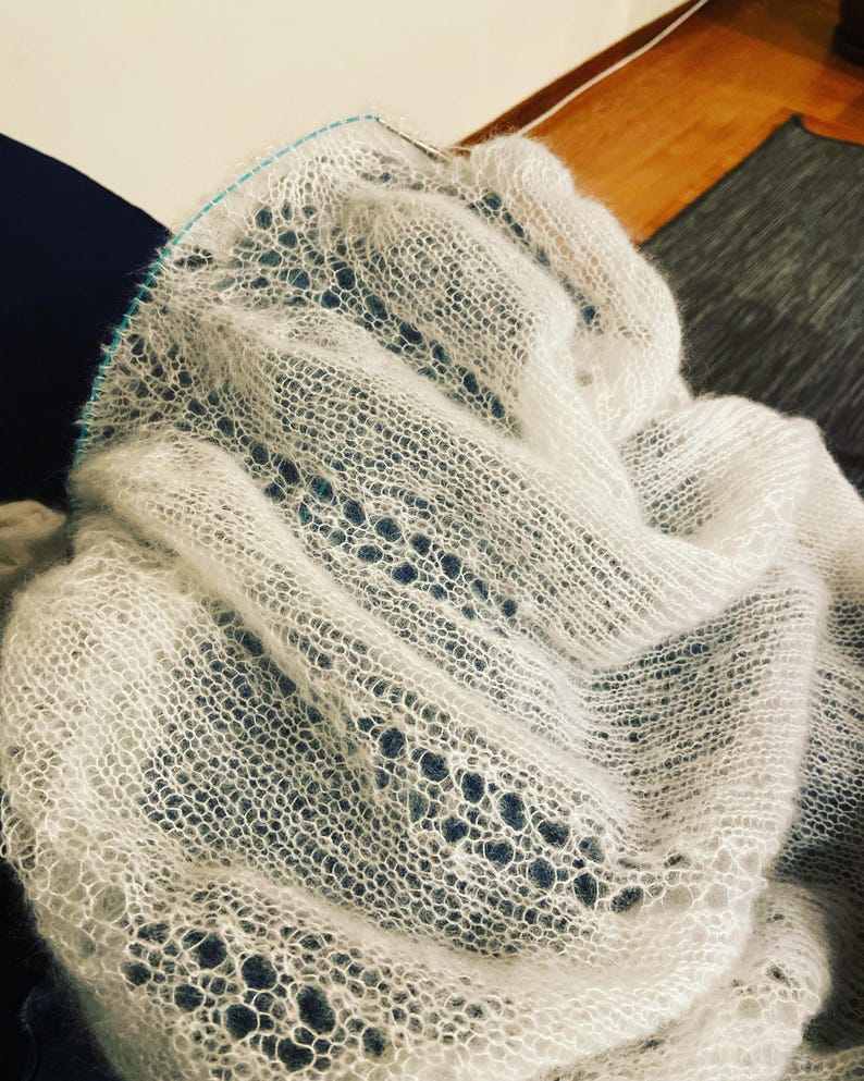 Elbert is a delicate, hand-knitted lace shawl made from soft, white mohair/silk yarn. The intricate lace pattern features openwork stitches, creating an airy and lightweight texture. The scalloped edges add elegance, making this piece perfect for formal occasions or as a cozy, stylish accessory. The natural light highlights the fine, haloed fibers, enhancing the ethereal quality of the knitwear. Ideal for those searching for luxury handmade shawls, bridal wraps, or heirloom-quality knitwear