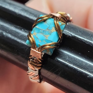 May include: A handmade wire-wrapped ring featuring a turquoise stone. The stone is a vibrant blue with gold flecks, set within a frame of copper and gold-toned wire. The ring's band is a mix of silver and copper wire.