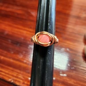 May include: A handmade ring with a peach-colored stone set in a copper wire wrap. The ring is displayed on a black ring holder against a warm-toned wooden surface. The wire is intricately woven around the stone, creating a unique, artistic design.