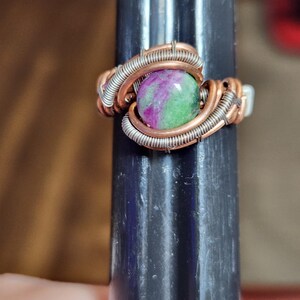 May include: A wire-wrapped ring featuring a round, polished ruby zoisite gemstone with vibrant magenta and green inclusions. The band is constructed from thick copper wire accented with thinner, tightly coiled silver-toned wire wrapping.